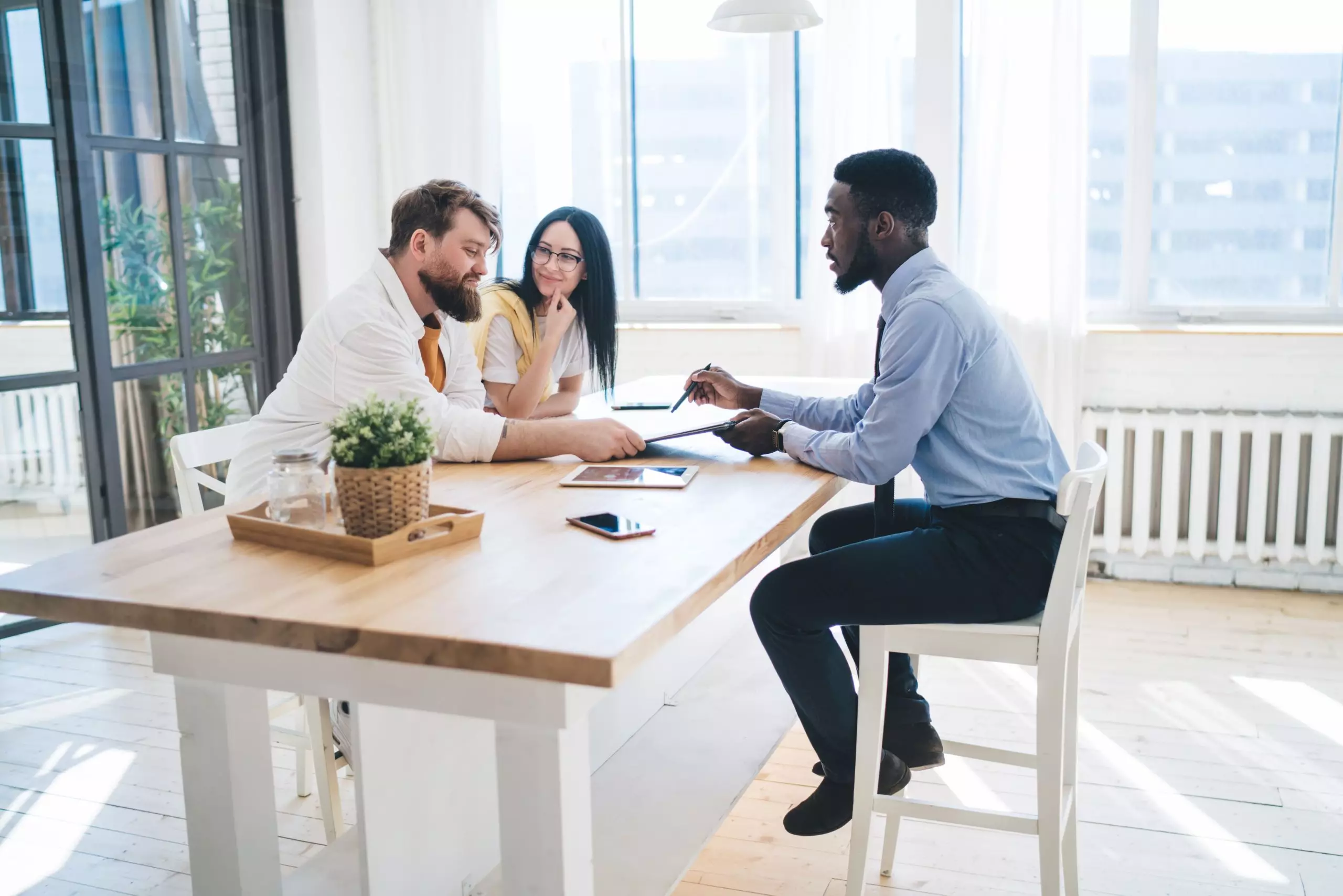 Side view of pleased couple sitting at table in bright room and smiling while presentable ethnic salesman offering apartment purchase contract